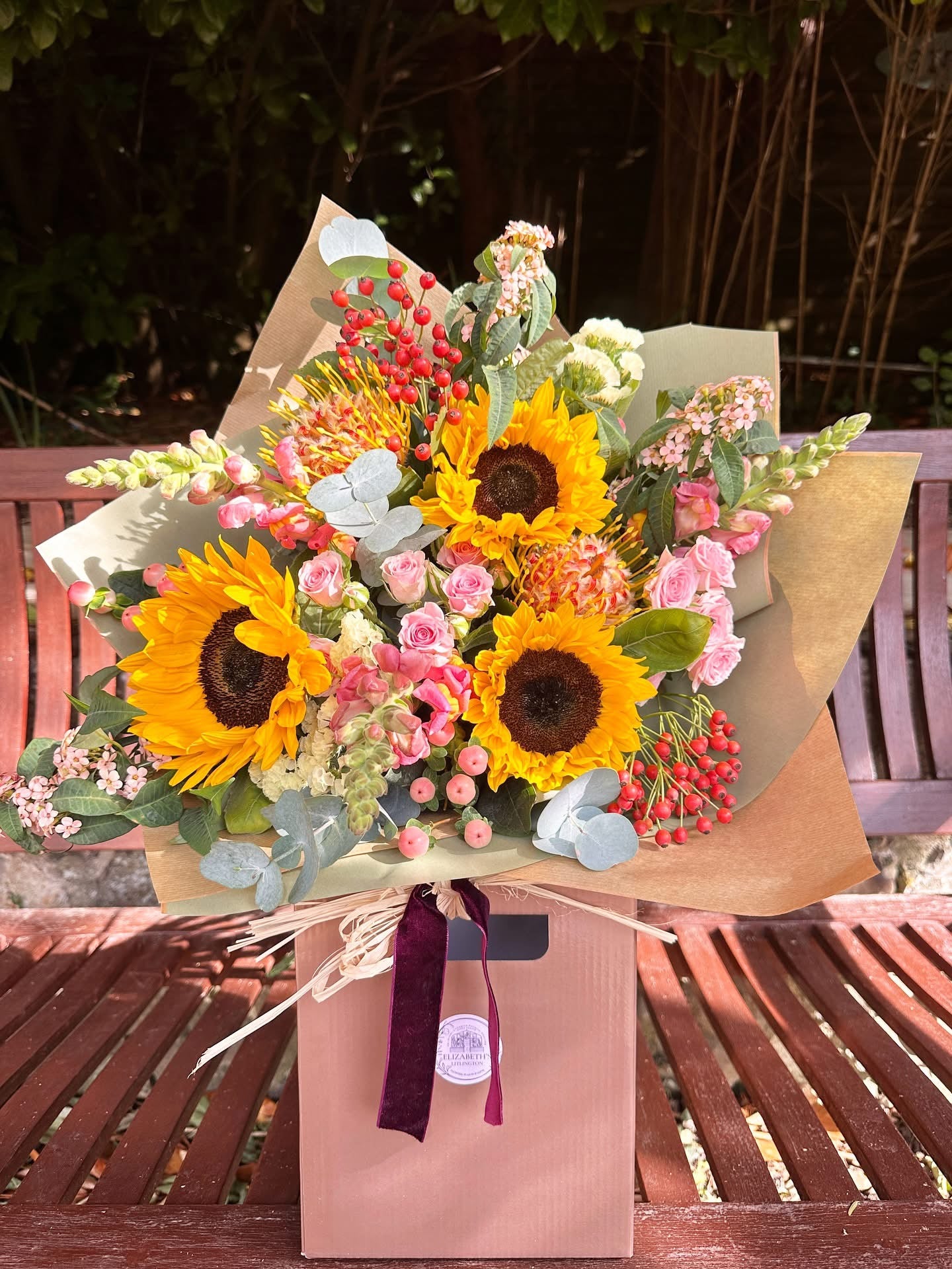 Bouquet of sunflowers and other flowers wrapped in brown paper with a pink gift bag on a wooden surface.